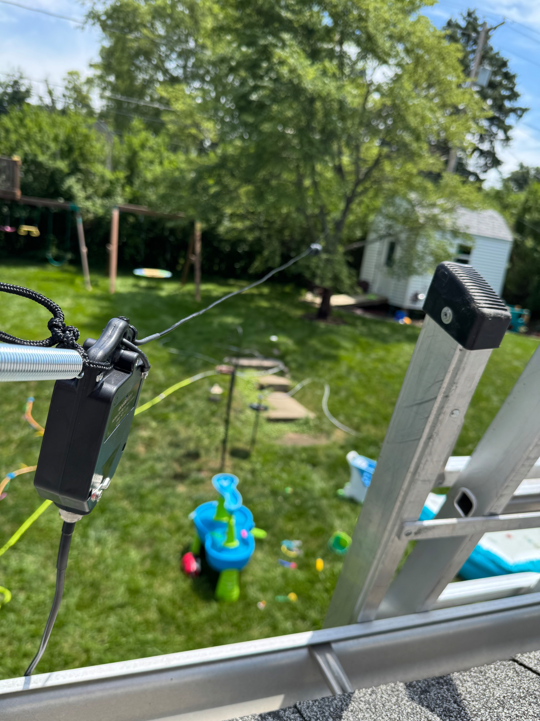 A close-up view from a roof shows a fishing rod setup with a grassy backyard, children's toys, and a small white shed in the background.